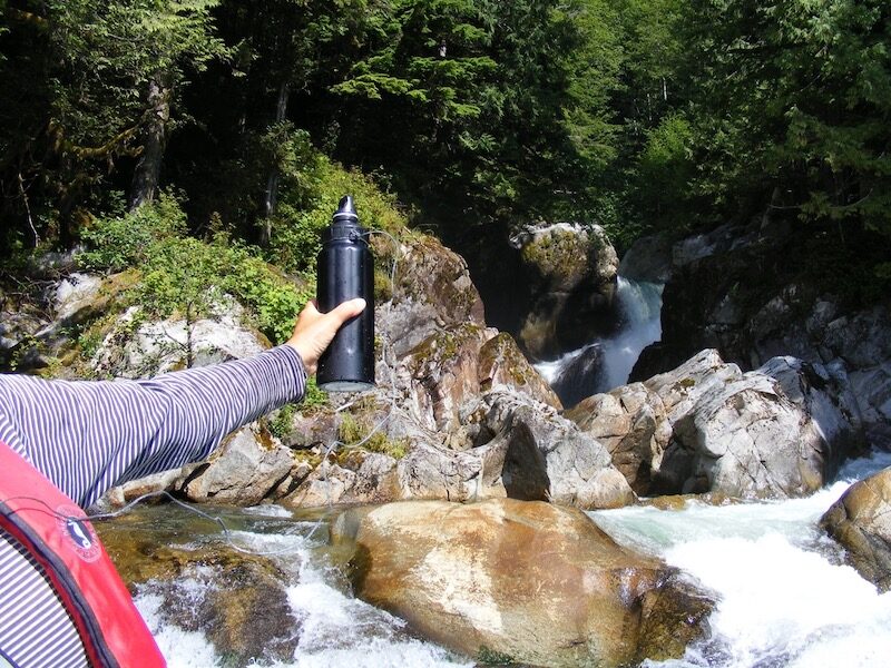 A hand holds out a black metal water bottle in front of the Paradise River.