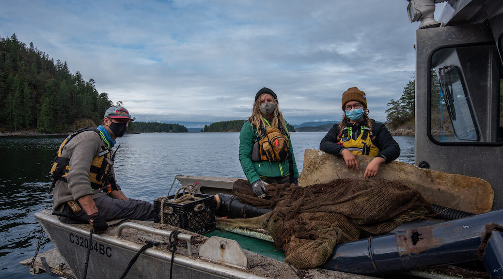 Three youth, all wearing masks and raingear, in a barge full of debris.