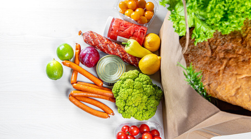 A paper bag full of groceries, surrounded by of fruits, vegetables, sausages and canned goods