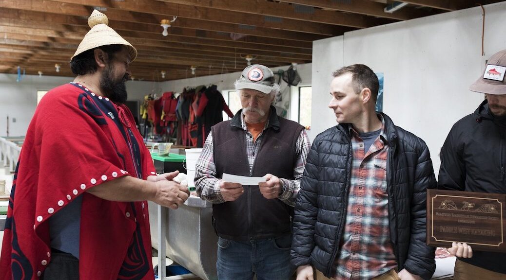 Indigenous man, in traditional dress, hands cheque to two men dressed in the casual gear of sports fishermen