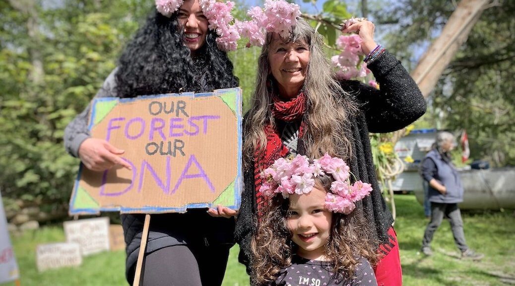 Two women and a girl with flowers in their hair. One of the women carries a sign saying 'Our forest, our DNA'