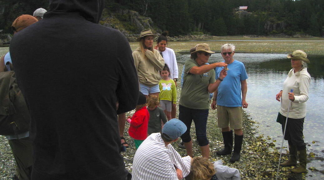woman standing beside a lagoon holding parts of a dead crab, a number of people look on