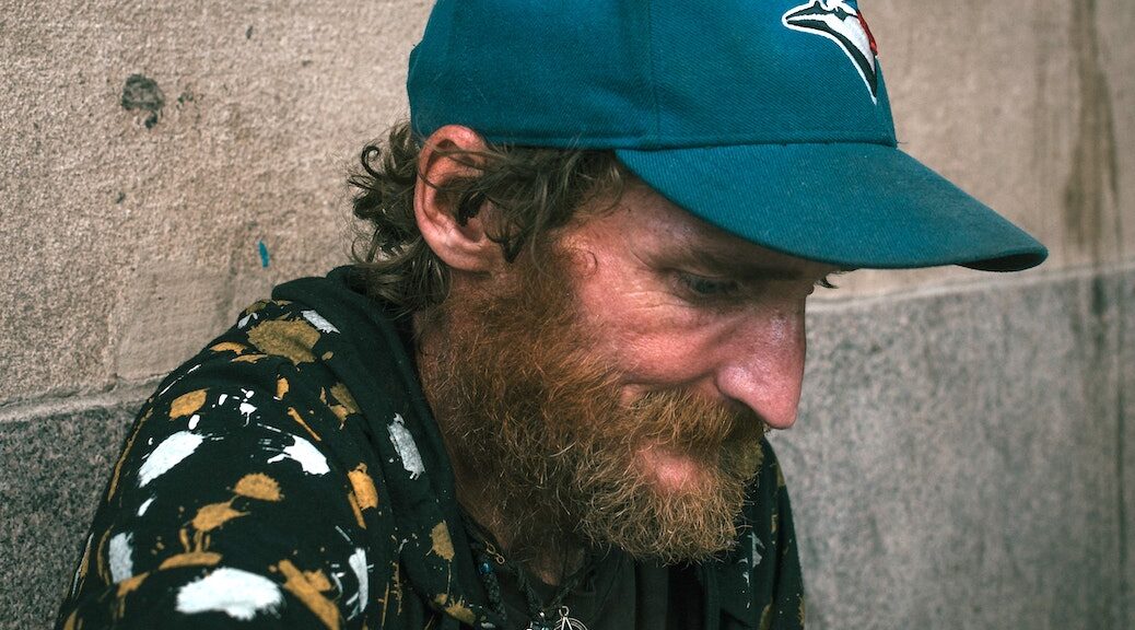 A homeless man, wearing a Toronto Bluejays cap, looks down