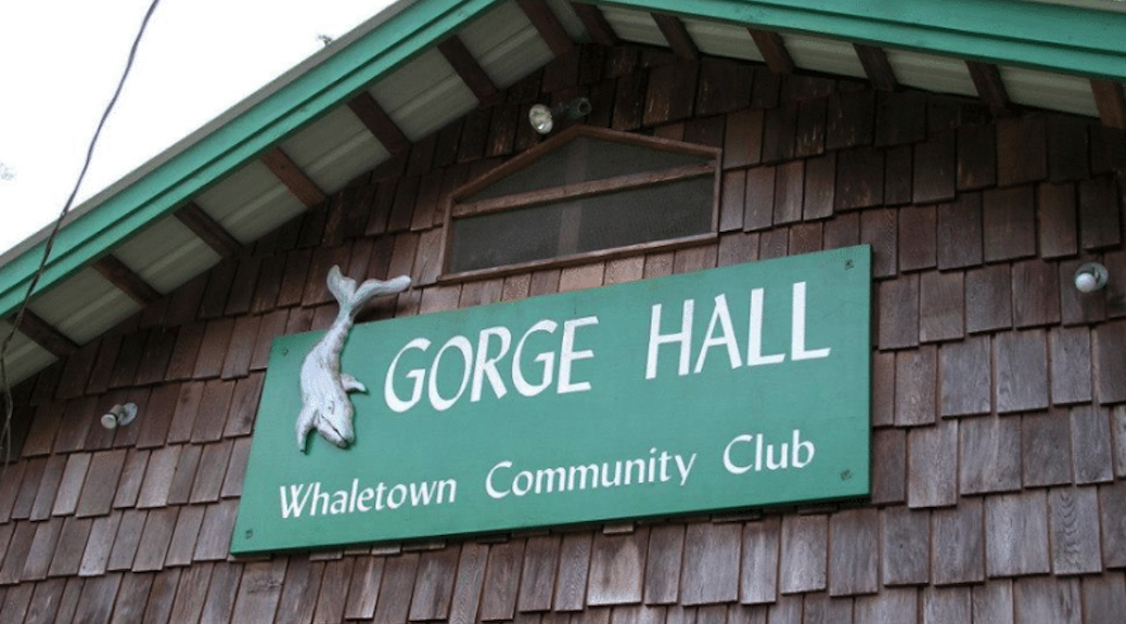 A green and white sign with the words Gorge Hall on a shake gable end with green facia boards. There is a sign