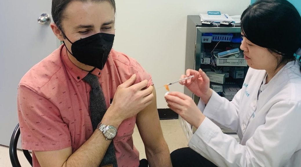 Seated man, with black mask on, getting a vaccination