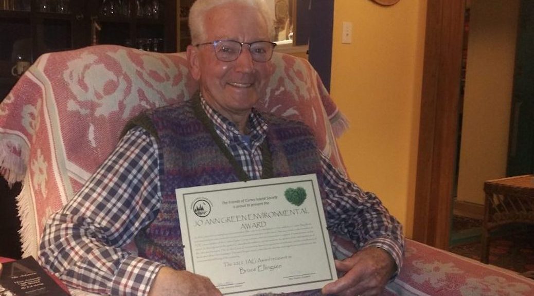 A smiling older man, sitting in an easy chair, holds a certificate.