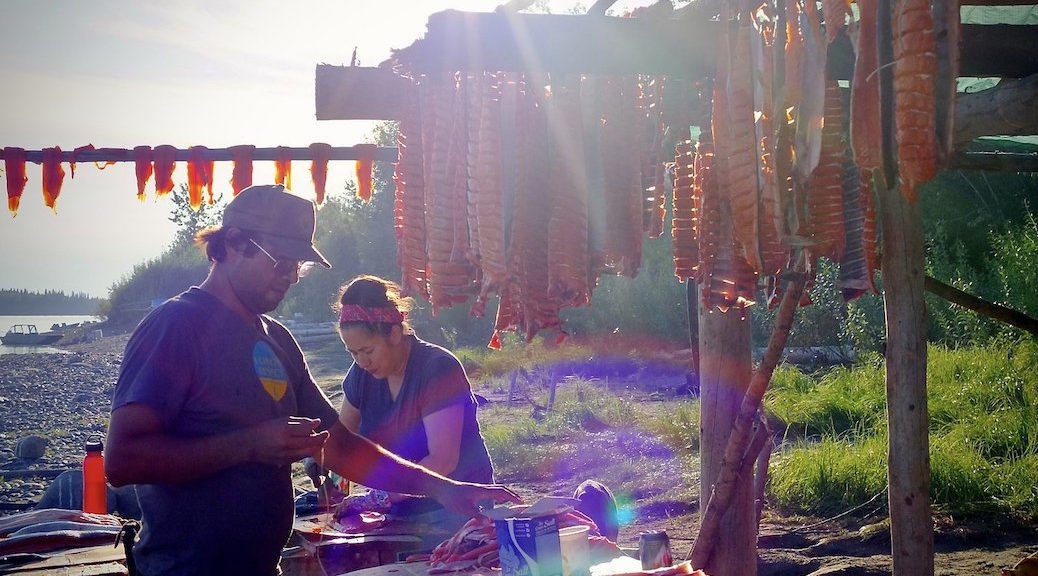 A sandy beach, with trees behind. An Indigenous man and woman stand at opposite sides of a table. Racks of drying salmon are around them.