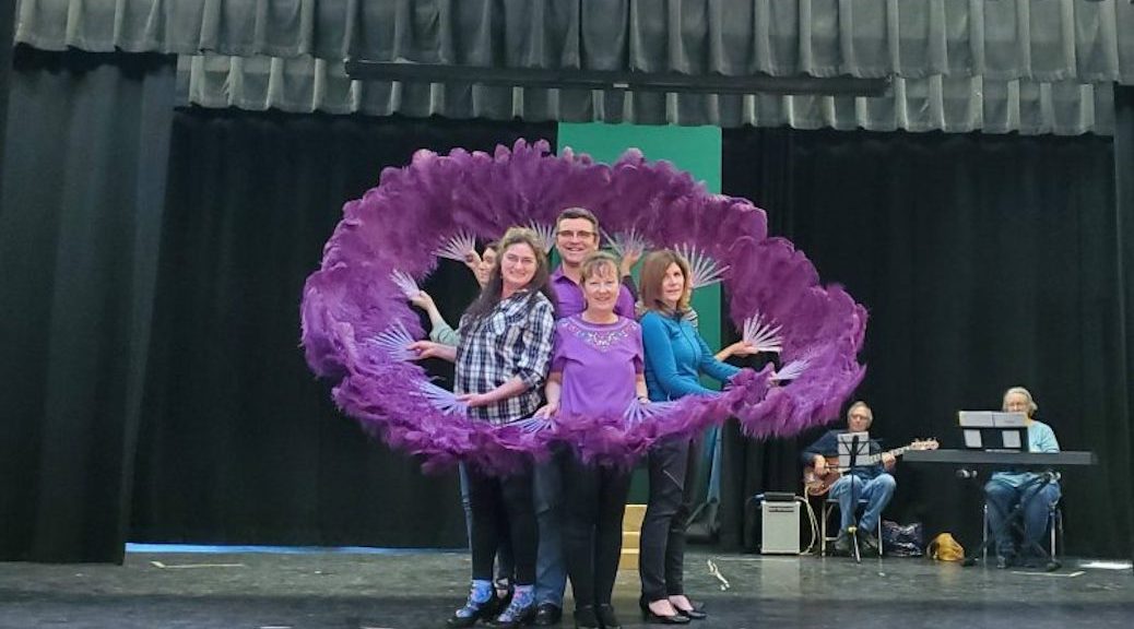 Three women and a man stand in the middle of an array of purple feathers