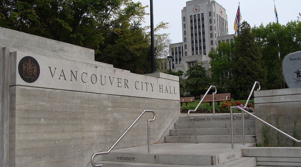 Steps running up beside a brick wall with the words 'Vancouver City Hall' at the top. The city hall rises in the background.
