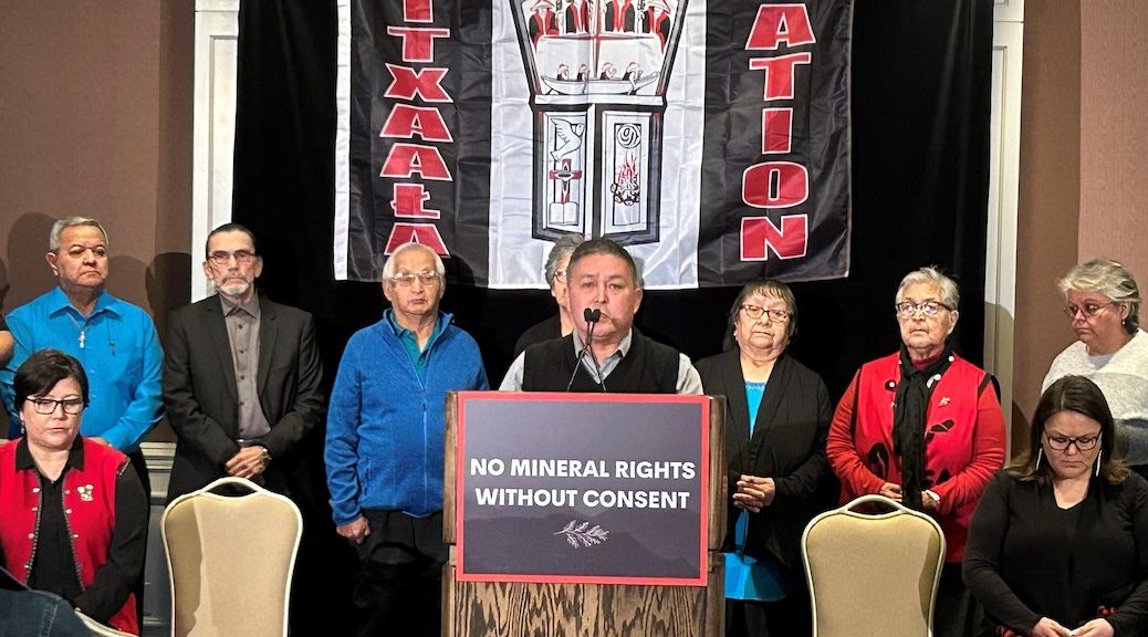 An Indigenous man speaks into a mic, seven other Indigenous people are grouped behind him.
