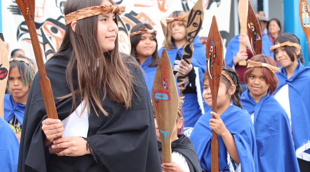 Children dressed in INdigenous costumes and carrying paddles smile for a photo op