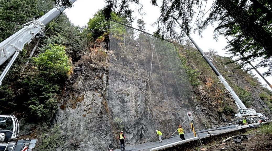 Two gigantic lifts have set a screen in front of a stone cliff, overlooking the highway where four highway workers are intently studying the pavement