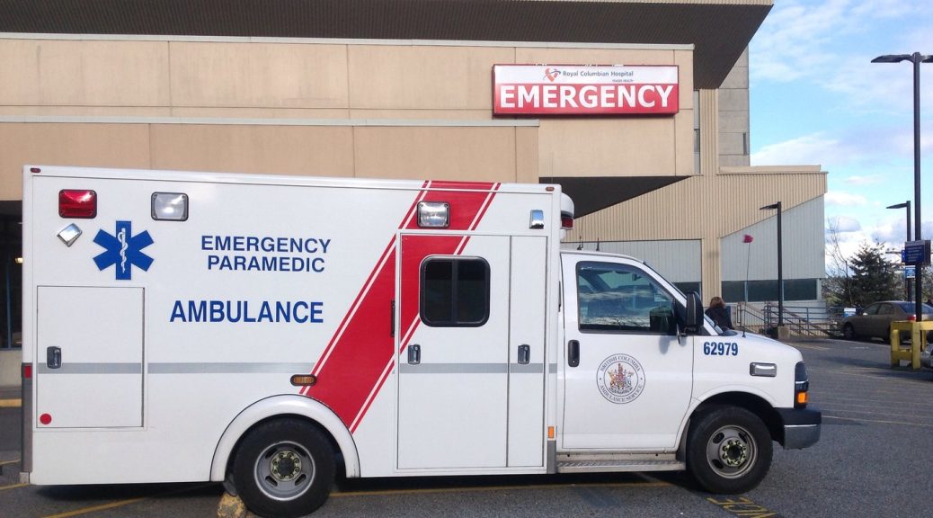 An ambulance, with the words 'Emergency Paramedic Ambulance' is parked beside the Emergency entrance of a hospital.