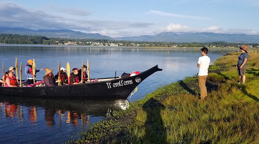 Two people stand on the shore as the canoe 'Tl-emtl'ems' brings INdigenous people to land