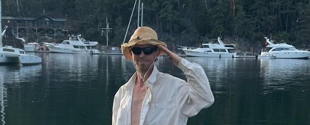 Man in a white sghirt and straw hat saluting to the camera. there are boats in a harbour behind him.