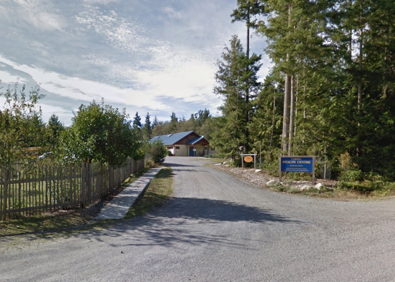 looking down a driveway towards a one story building with a turquooise roof. A sign says 'Cortes Health Centre'