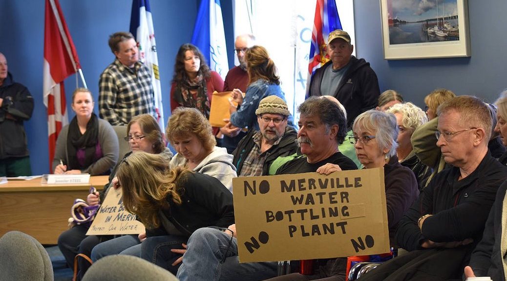 Sitted man, in a croweded room, displays d=sign saying 'No Merville Water Bottling Plant'