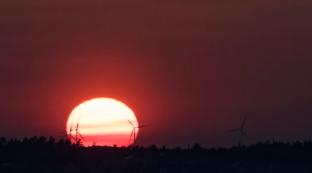 Wind turbines are framed by the sun setting in a red (smoke filled) sky.