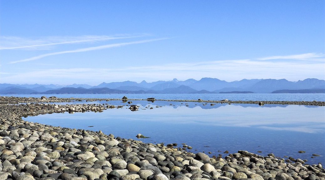 Looking across the waters from a rocky beach to distant islands. The icture is dominated by blue sky and ocean