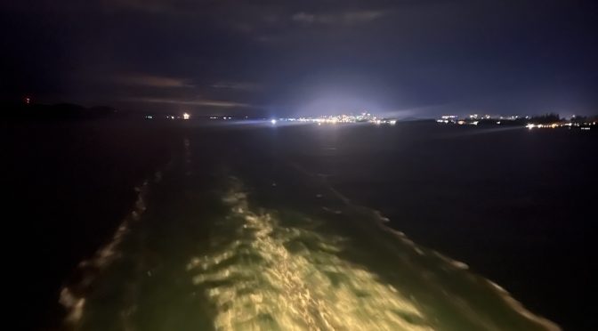 Looking back over thw wake of a ship to city lights in a dark sky