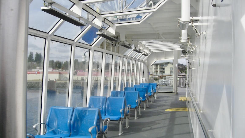 A glassed in area on the upper deck of a ferry. Pairs of blue seats butt up against the window