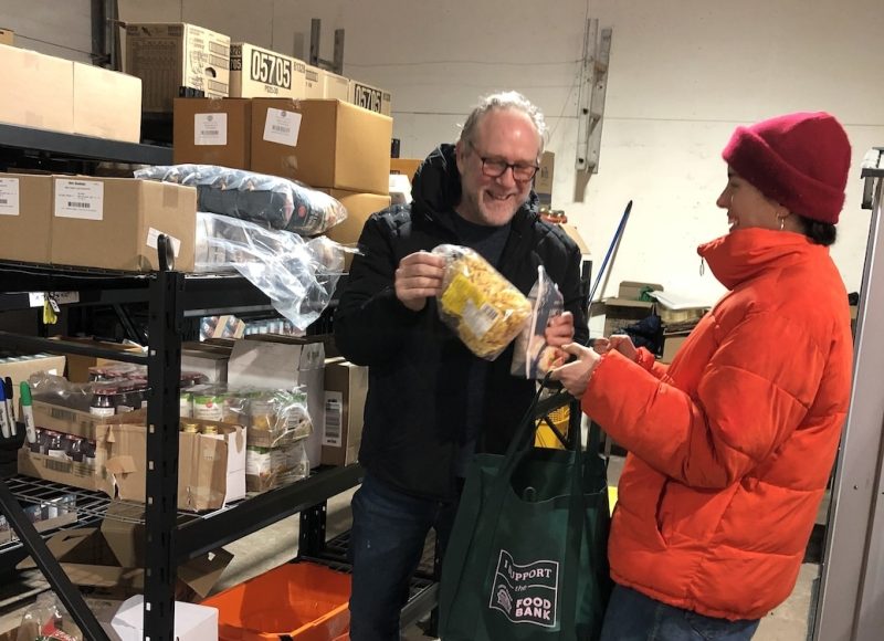 A man and a woman standing in a wharehouse full of fordstuffs. They are filling a bag with groceries.