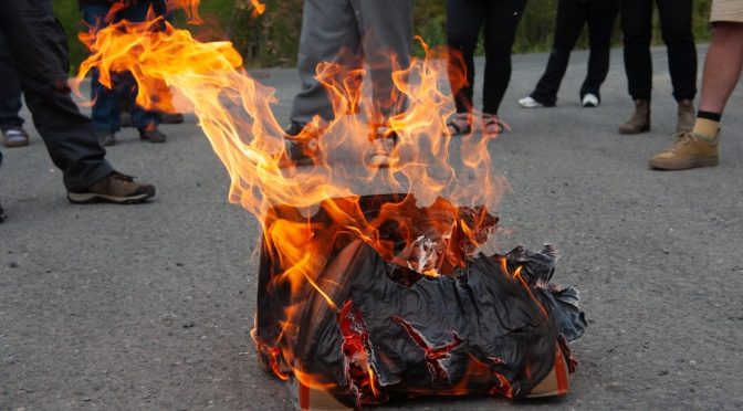 Burning briefcase on pavement surrounded by people's legs