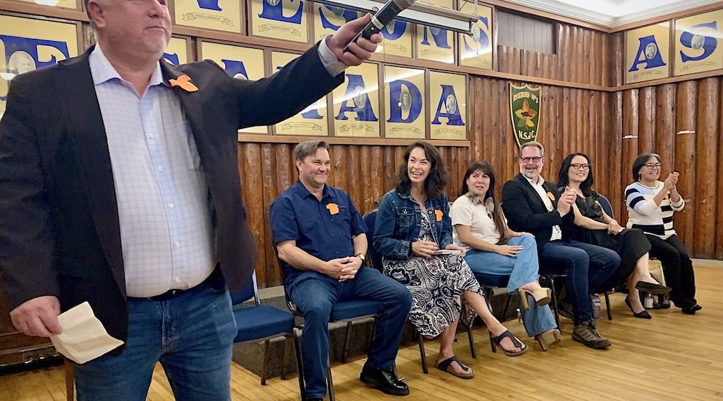 Man in front of hall holds mic towards audience. half a dozen other men and women to his left look on