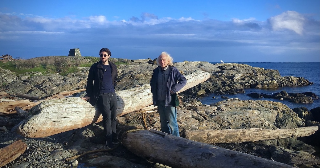 A young man and older man stand amidst drift logs on a beach