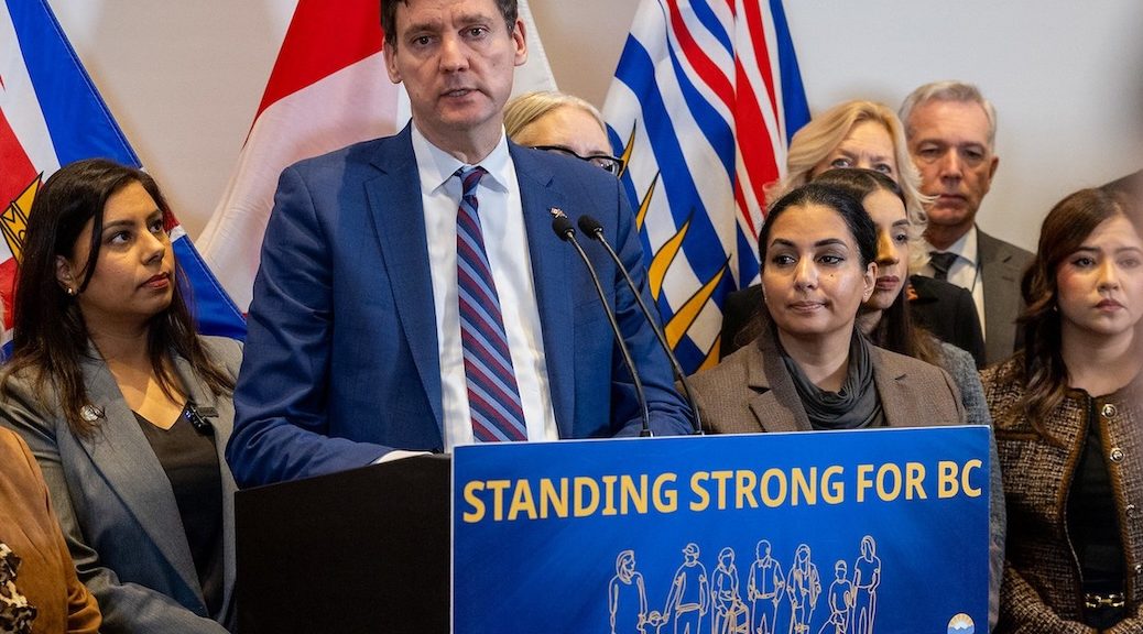 A very tired looking Premiere David stands at a podium. A number of people stand behind him.