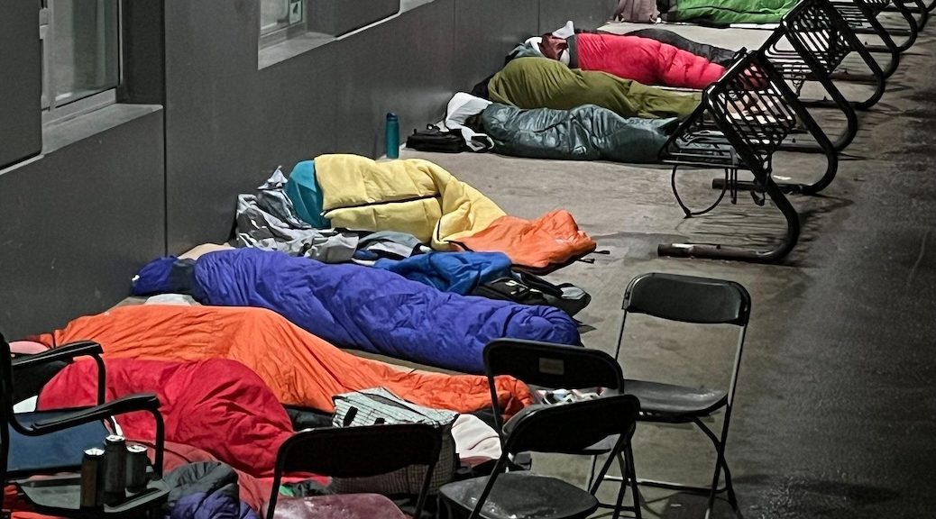 A line of people, whose faces are covered, in sleeping bags on a sidewalk