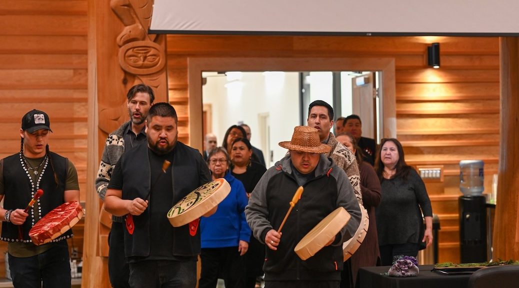 Two drummers lead a group of indigenous people into a room