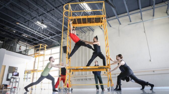 Two dancers on a scaffolding, which other dancers are holding