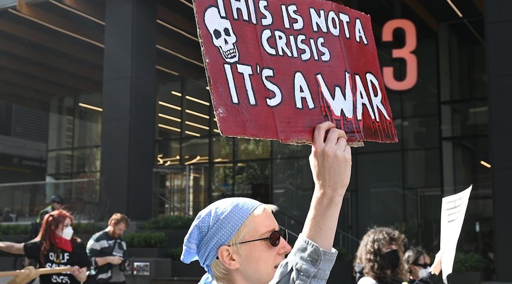 A woman, in the midst of a protes heading down the street, holds up a sign that says 'This is not a crisis, it is a war."
