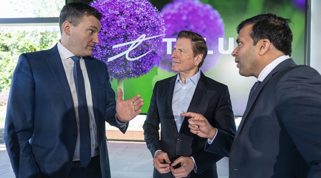 Three men having an intense conversation in front of a large TELUS sign. One is pointing his finger, another is gesturing with an open hand