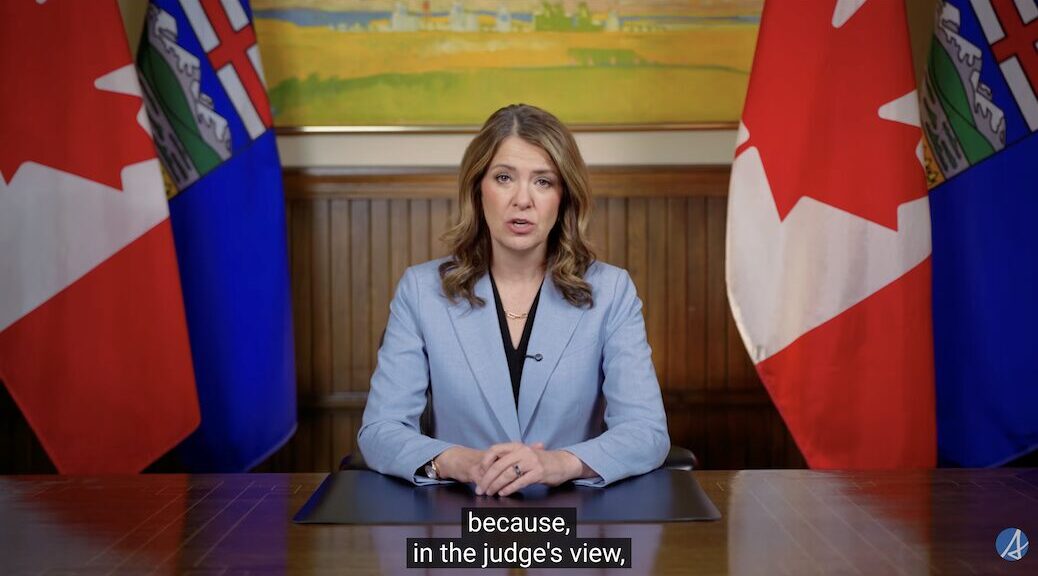 Woman with hands clasped, sitting in front of Albertan and Canadian flags, addressing the province