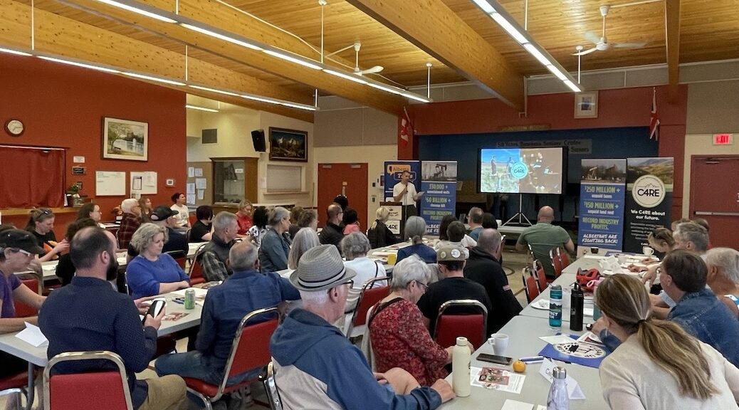 A room full of people sitting at long tables listen to a speaker
