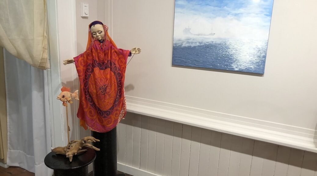 Female puppet with outstretched arms, dressed in red, beside a seascape full of light and mist with the faint outline of a distant freighter