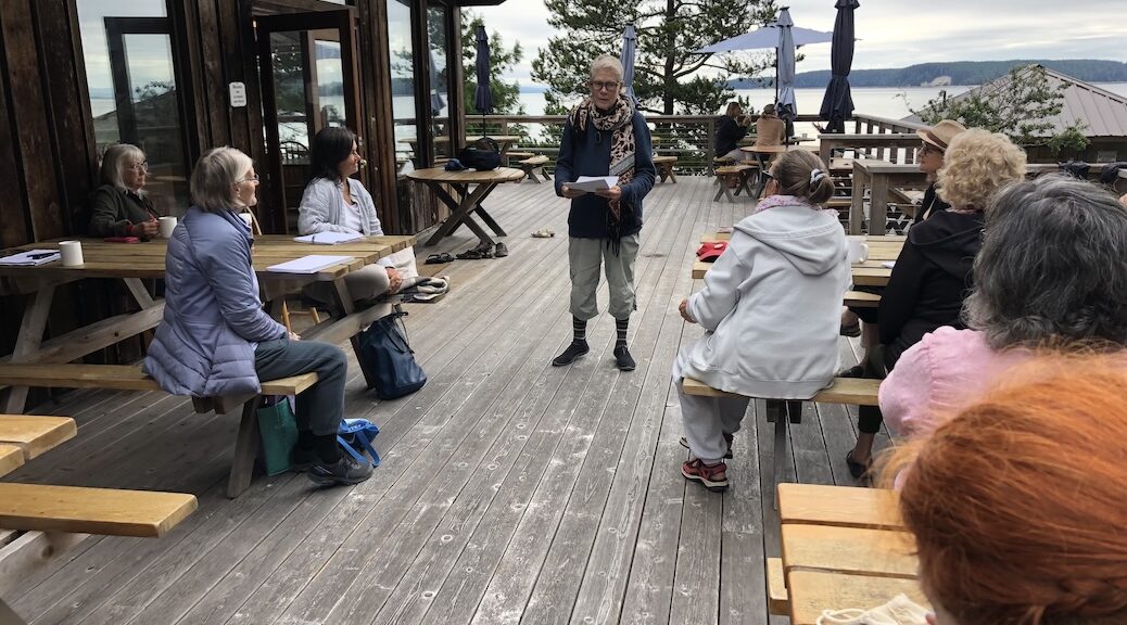 Woman with a sheet of paper in her hands standing on an open deck. Eight wother women are intenting listening to her.