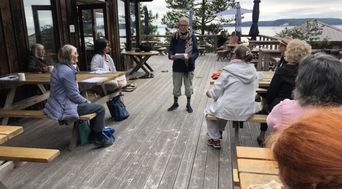 Woman with a sheet of paper in her hands standing on an open deck. Eight wother women are intenting listening to her.